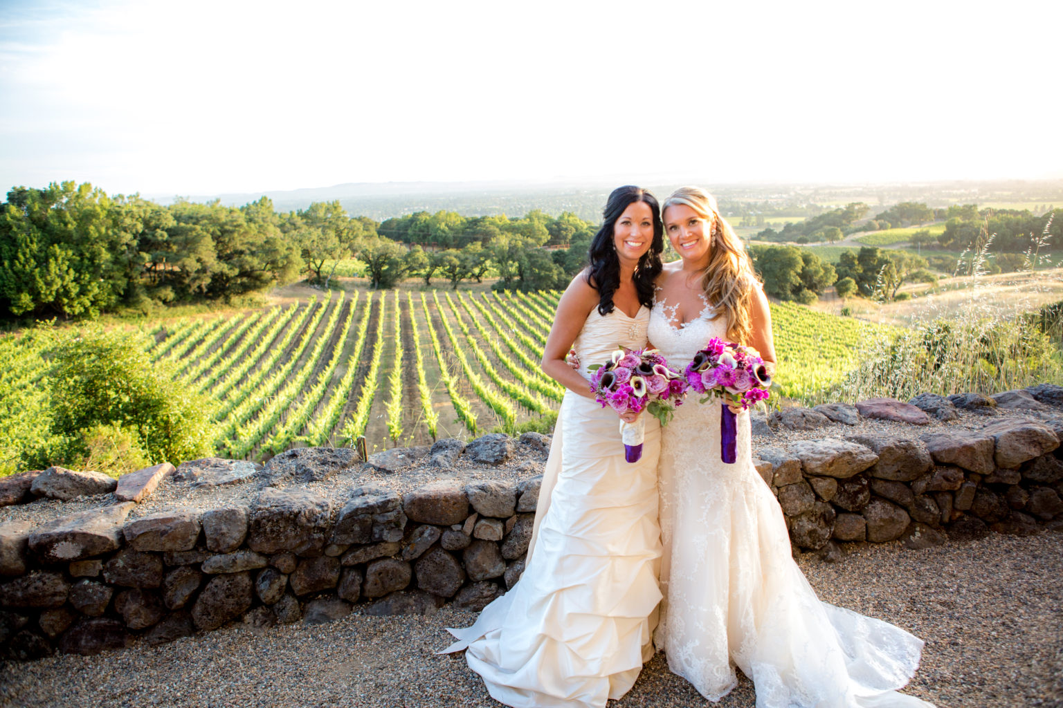 Brides smiling together in front of vineyards and oak trees below at Paradise Ridge winery
