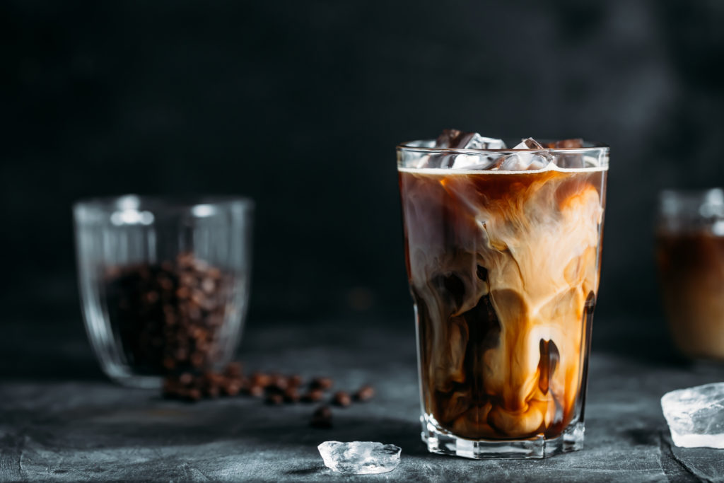 Ice coffee in a tall glass with cream poured over and coffee beans on dark gray background.
