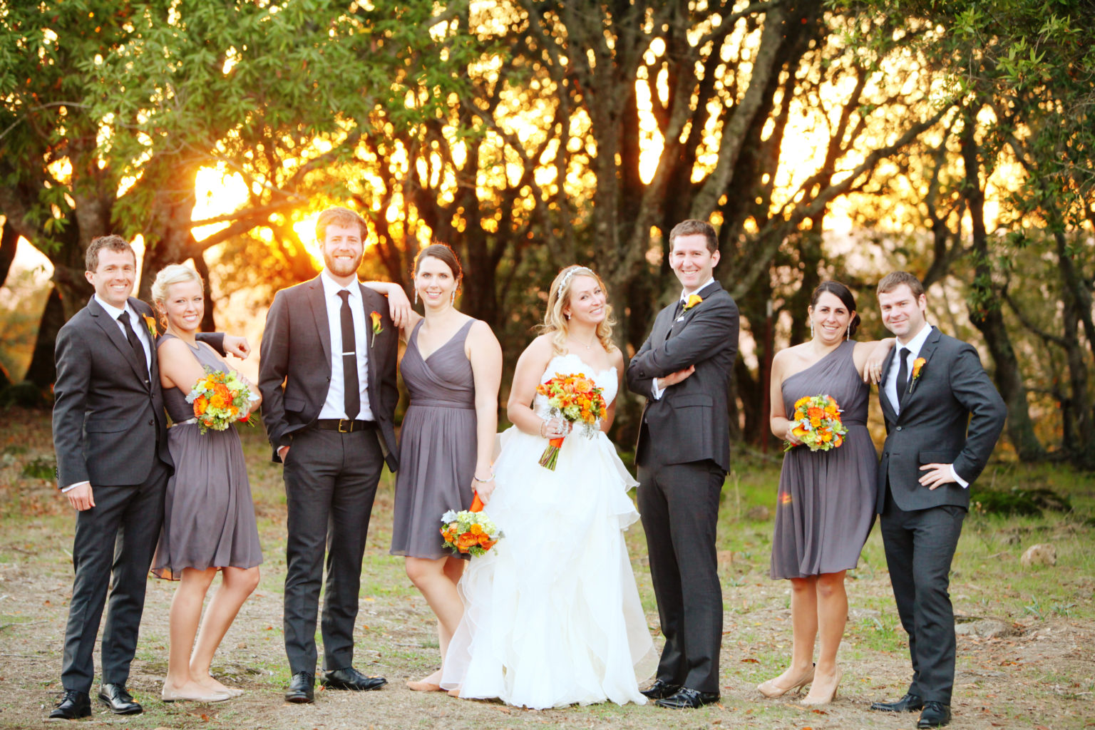 Wedding party smiling for photos in front of oak trees with sunset behind at Paradise Ridge winery